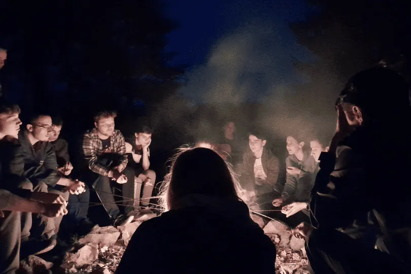Skupina mladých lidí v noci opéká špekáčky u ohně.