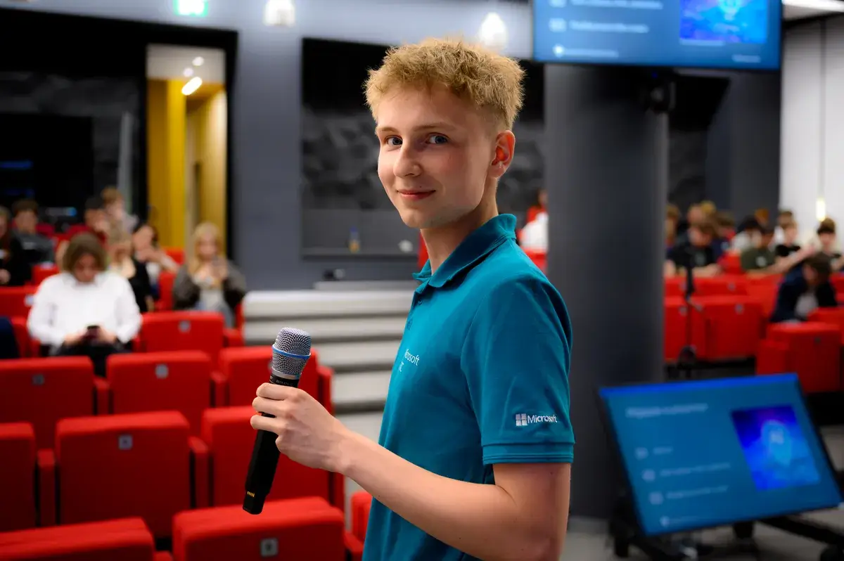 Student v tričku Microsoft s mikrofonem v ruce v přednáškovém sále plném lidí.